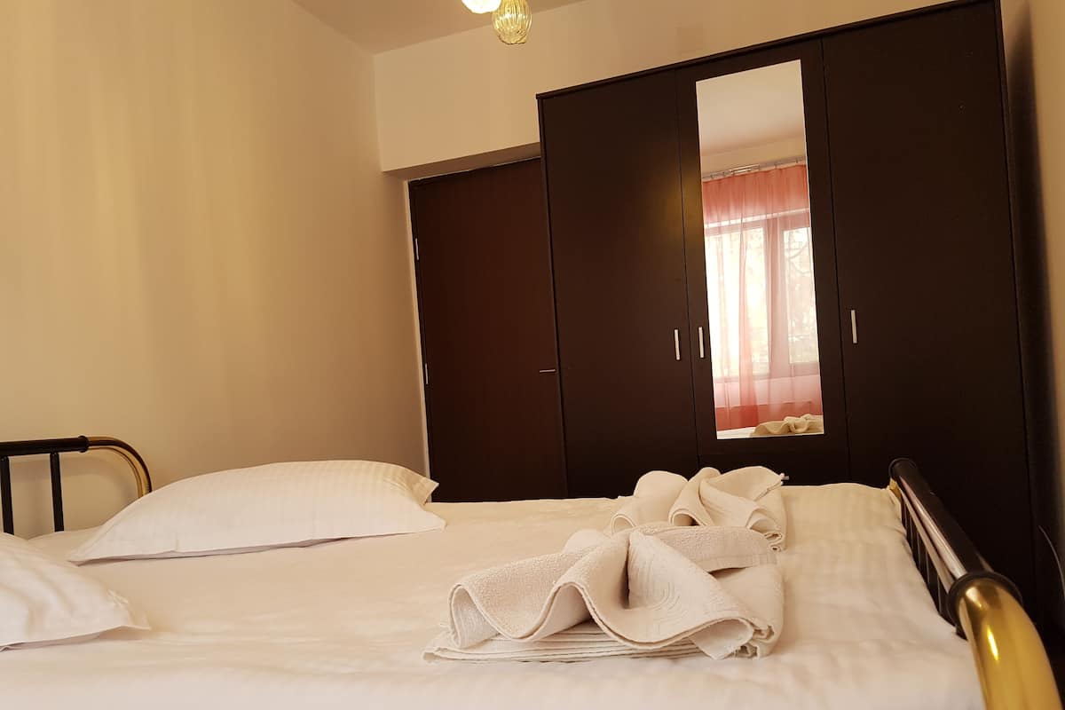 A neatly made bed with white linens and a folded towel, a dark wooden wardrobe, and a mirror reflecting a window with curtains.