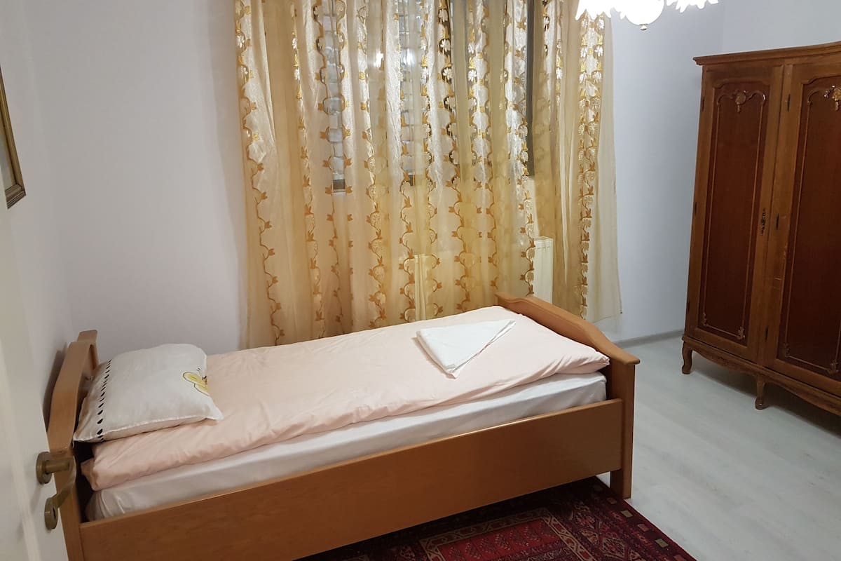 A single bed with a wooden frame, a white pillow, and a folded white towel. There is a wooden wardrobe to the right and a patterned rug on the floor.