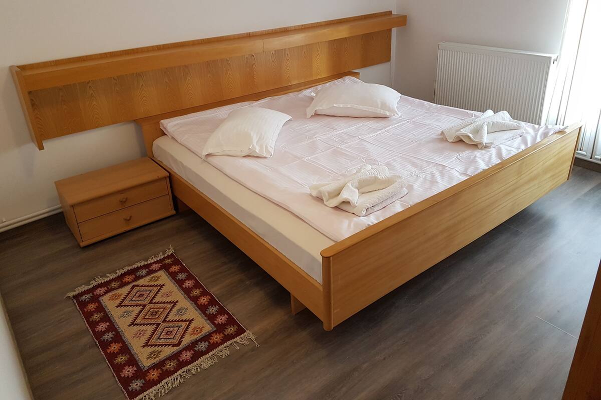 A neatly made bed with a wooden headboard, a small bedside table, and a patterned rug on a wooden floor.