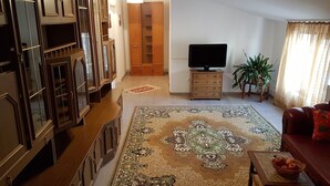 Een woonkamer met een grote houten kast, een televisie op een houten plank, een kamerbreed tapijt, een bank en een klein tafeltje met een mandje.