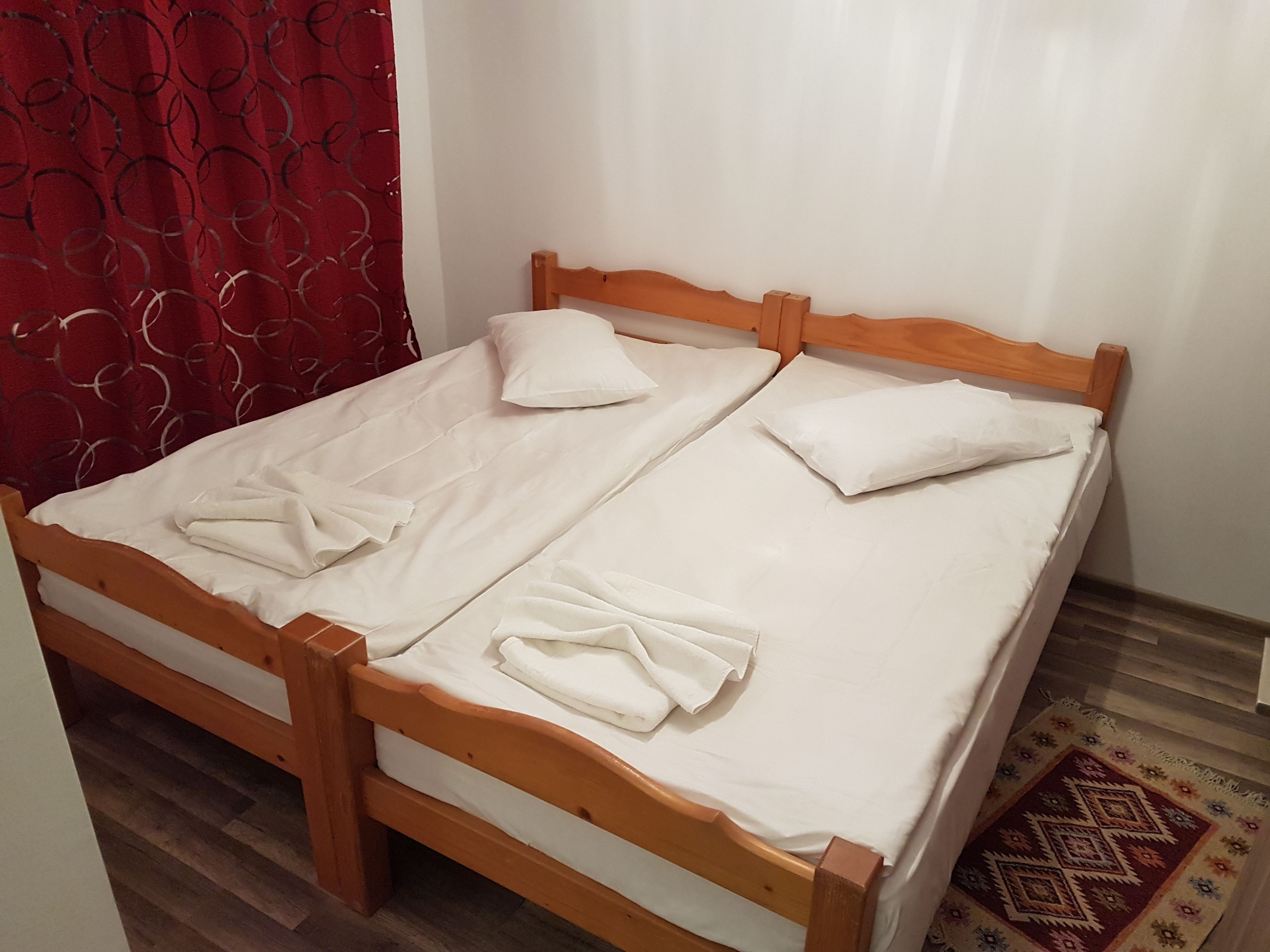 Cama doble con cabeceros de madera y ropa de cama blanca, cortina roja estampada y alfombra con diseño en el suelo.