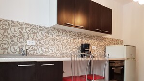 Een compacte keuken met donkere kastjes, een decoratieve spatwand, een koffiezetapparaat, een koelkast en een metalen stoel met een rode kussen.