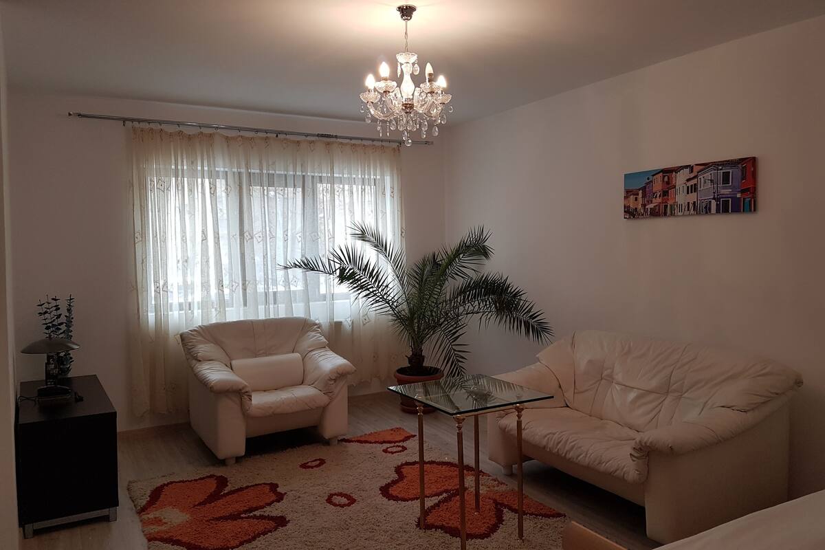 A living room with a beige sofa, a glass coffee table, a potted plant, a chandelier, and a painting on the wall.