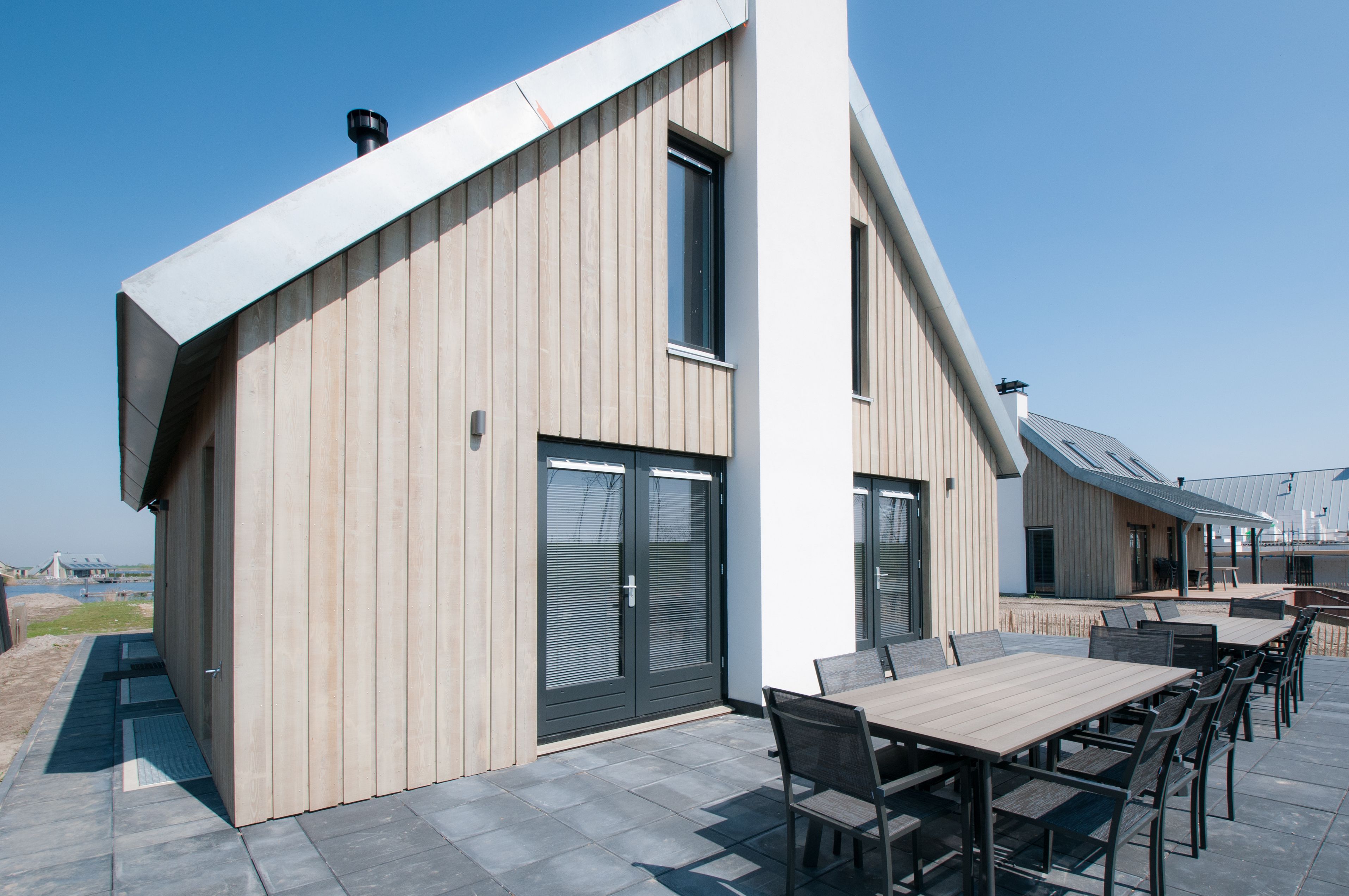 Een modern gebouw met een schuin dak, grote ramen en een terras met tafels en stoelen.