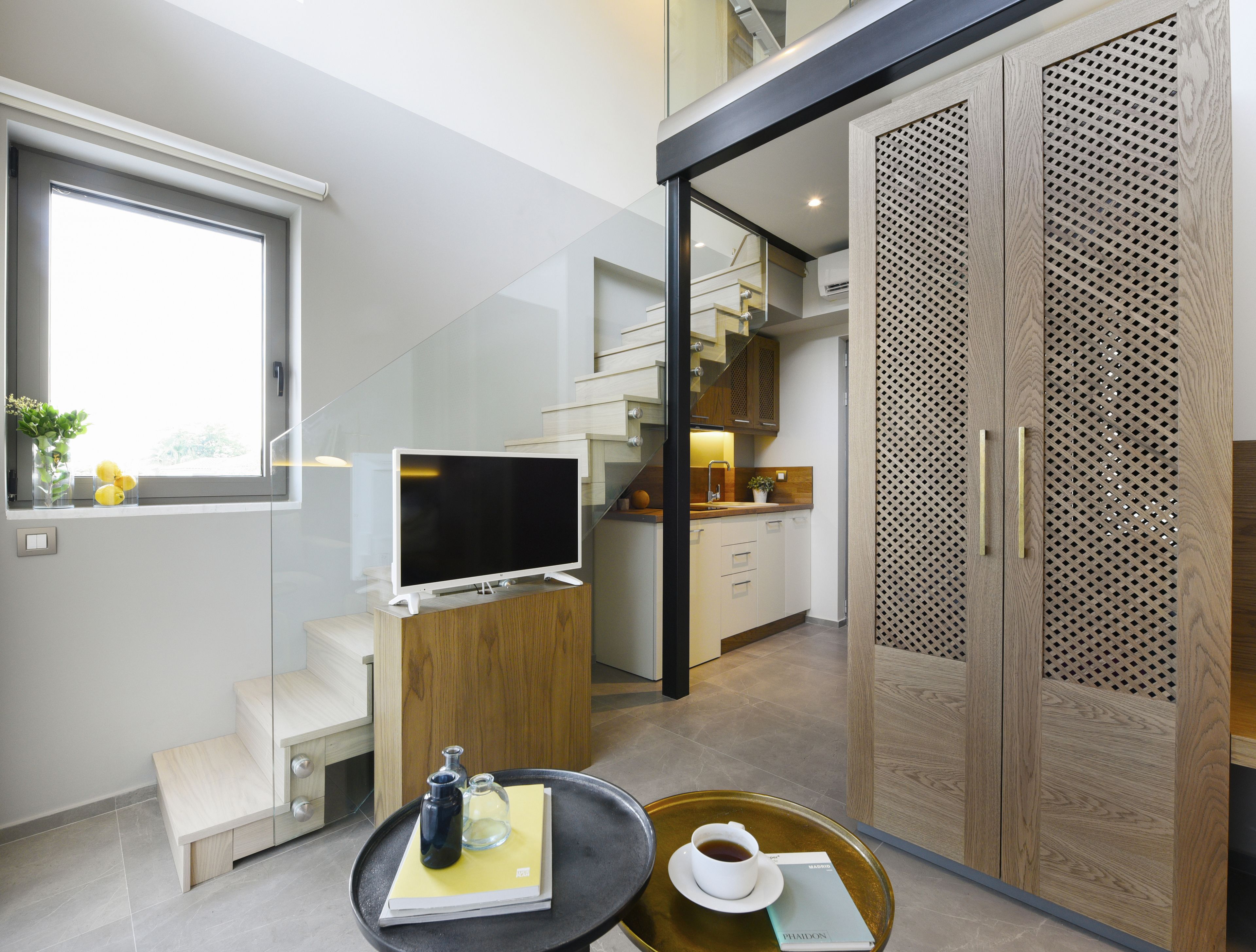 A modern living room with a flat-screen TV, a coffee table with a book and a cup, and a view of the kitchen and staircase.