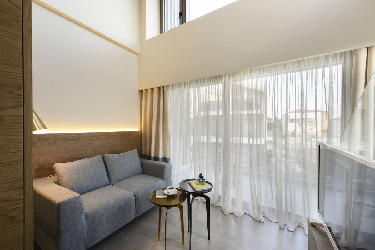 Una sala moderna con un sofá gris, una mesita redonda con un libro y una taza, y cortinas translúcidas.