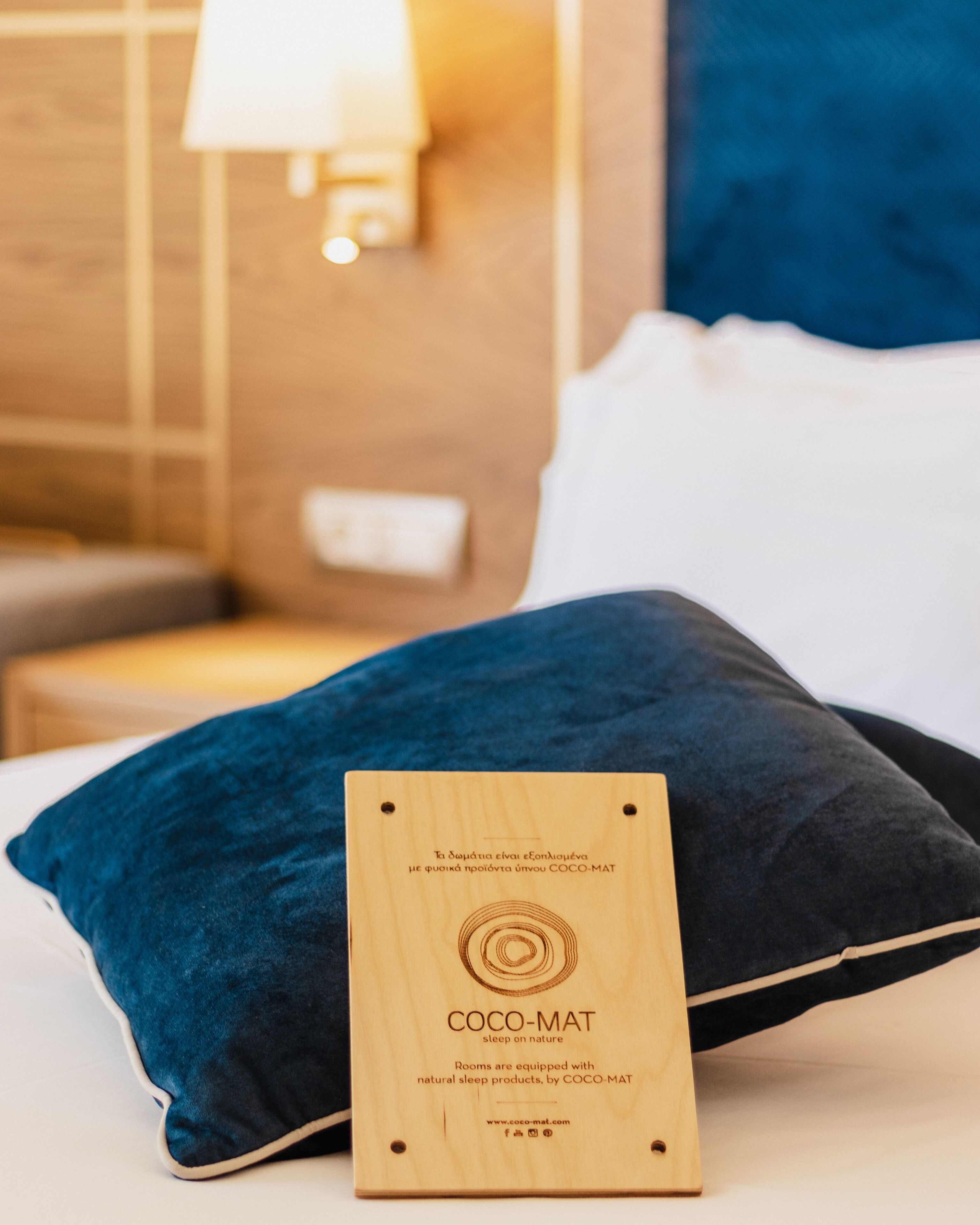 Una cama con una almohada azul oscuro y una placa de madera con la inscripción "COCO-MAT sleep on nature" en una habitación de hotel.