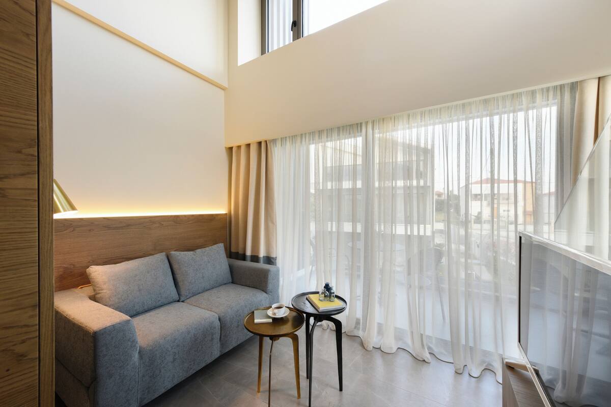 Una sala moderna con un sofá gris, una mesita redonda con un libro y una taza, y cortinas translúcidas.