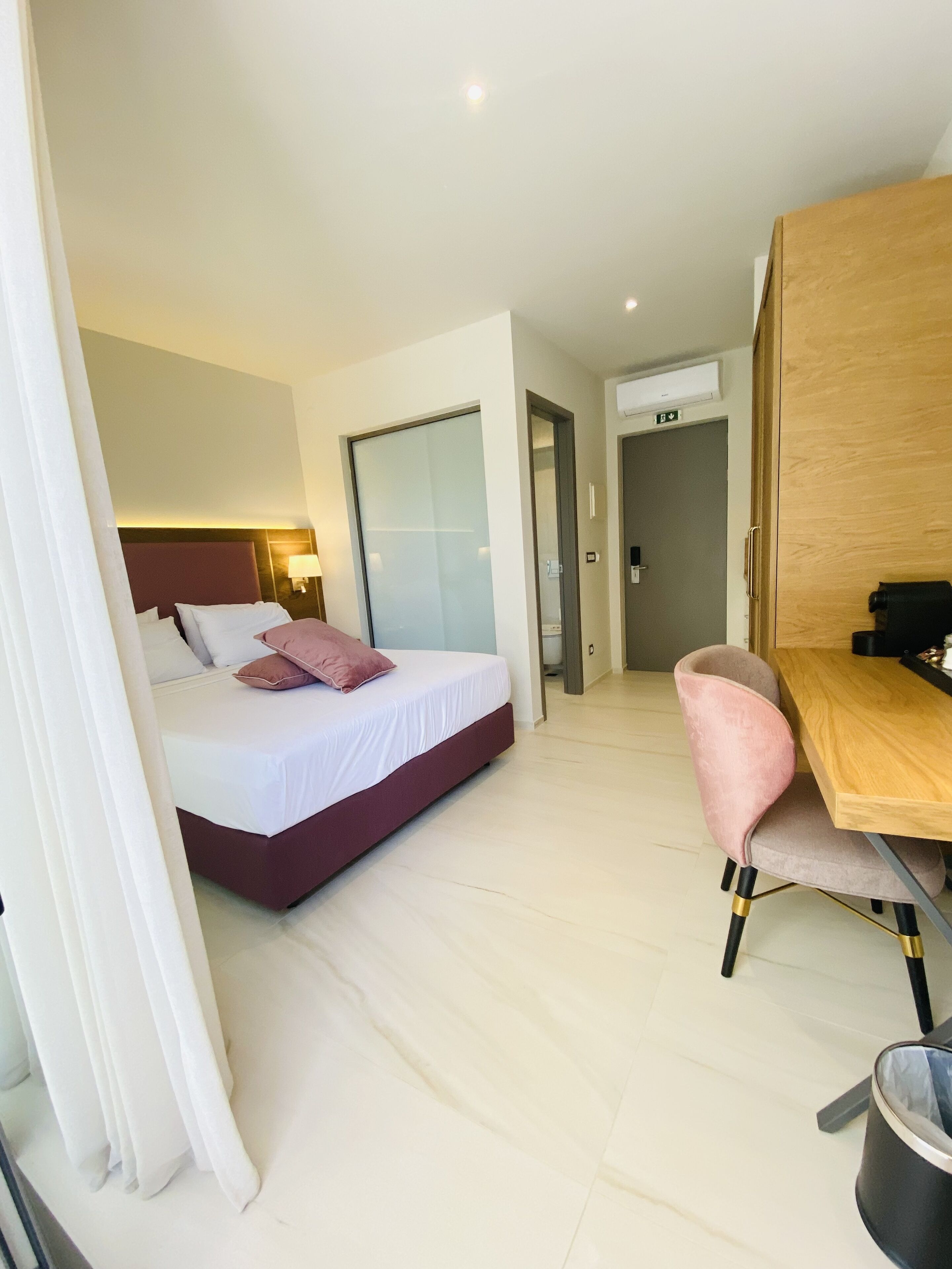A hotel room with a bed, a desk, and a chair. There is a door leading to another room and a trash can.