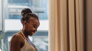 A person standing by a window with curtains, holding a cup. - Serenity Suites (Nikiti)