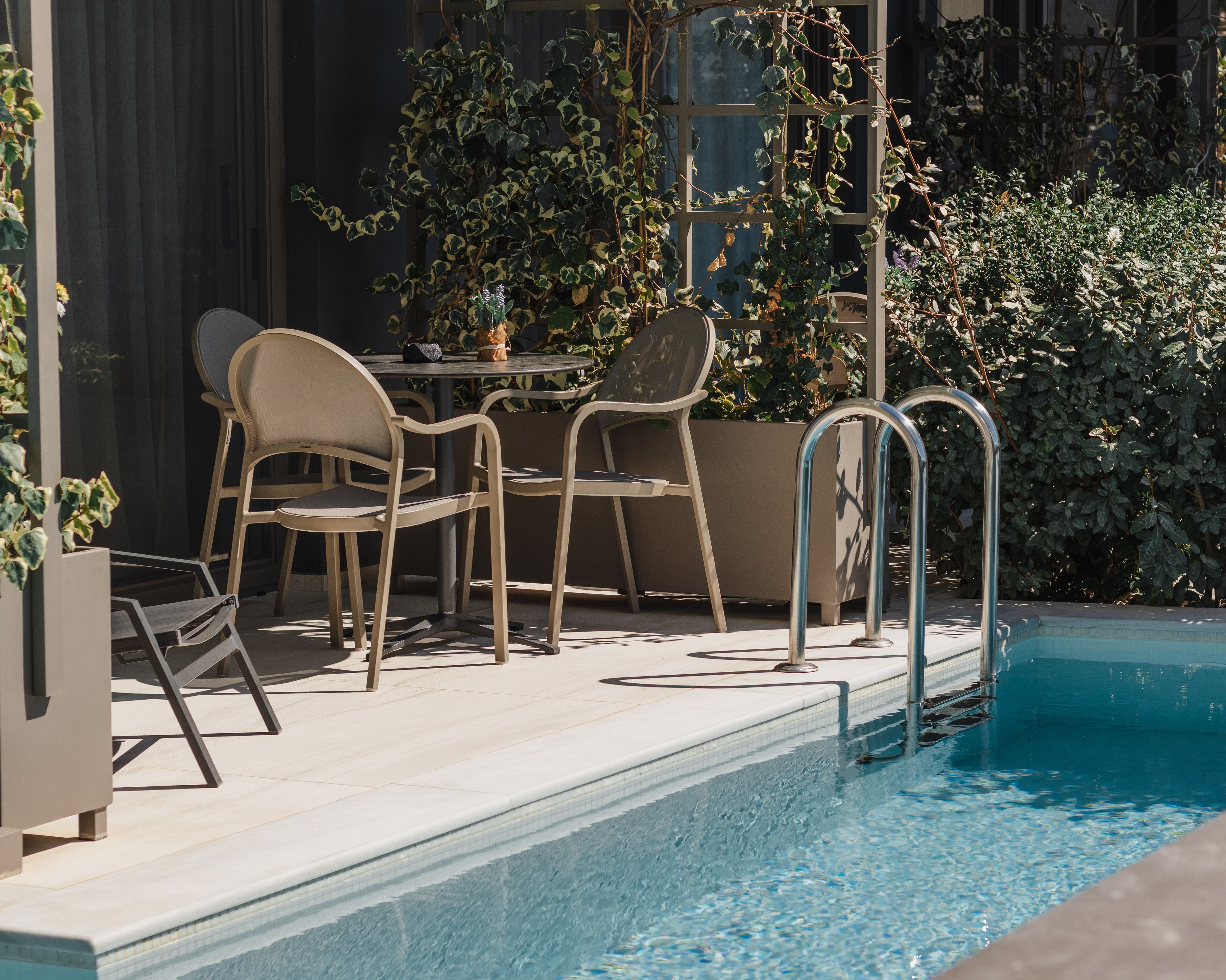 A poolside area with a table and chairs, a metal pool ladder, and surrounding greenery.