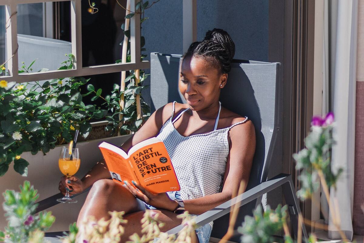 Una persona sentada en una silla leyendo un libro al aire libre, con plantas y una ventana de fondo.