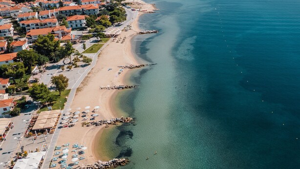 An aerial view of a coastal town with a beach, clear waters, and red-roofed buildings. - Serenity Suites (Nikiti)