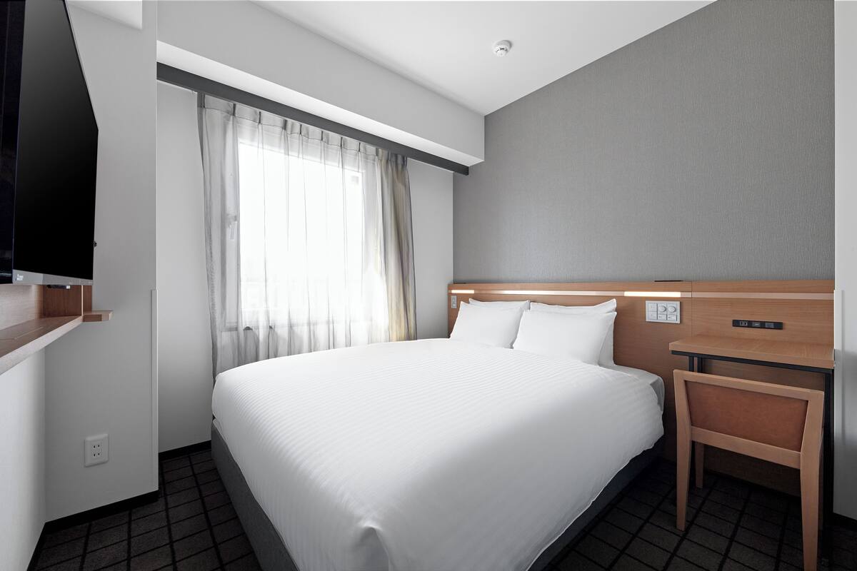 Une chambre d’hôtel avec un grand lit, une table de chevet en bois, un téléviseur à écran plat et une fenêtre ornée de rideaux légers.