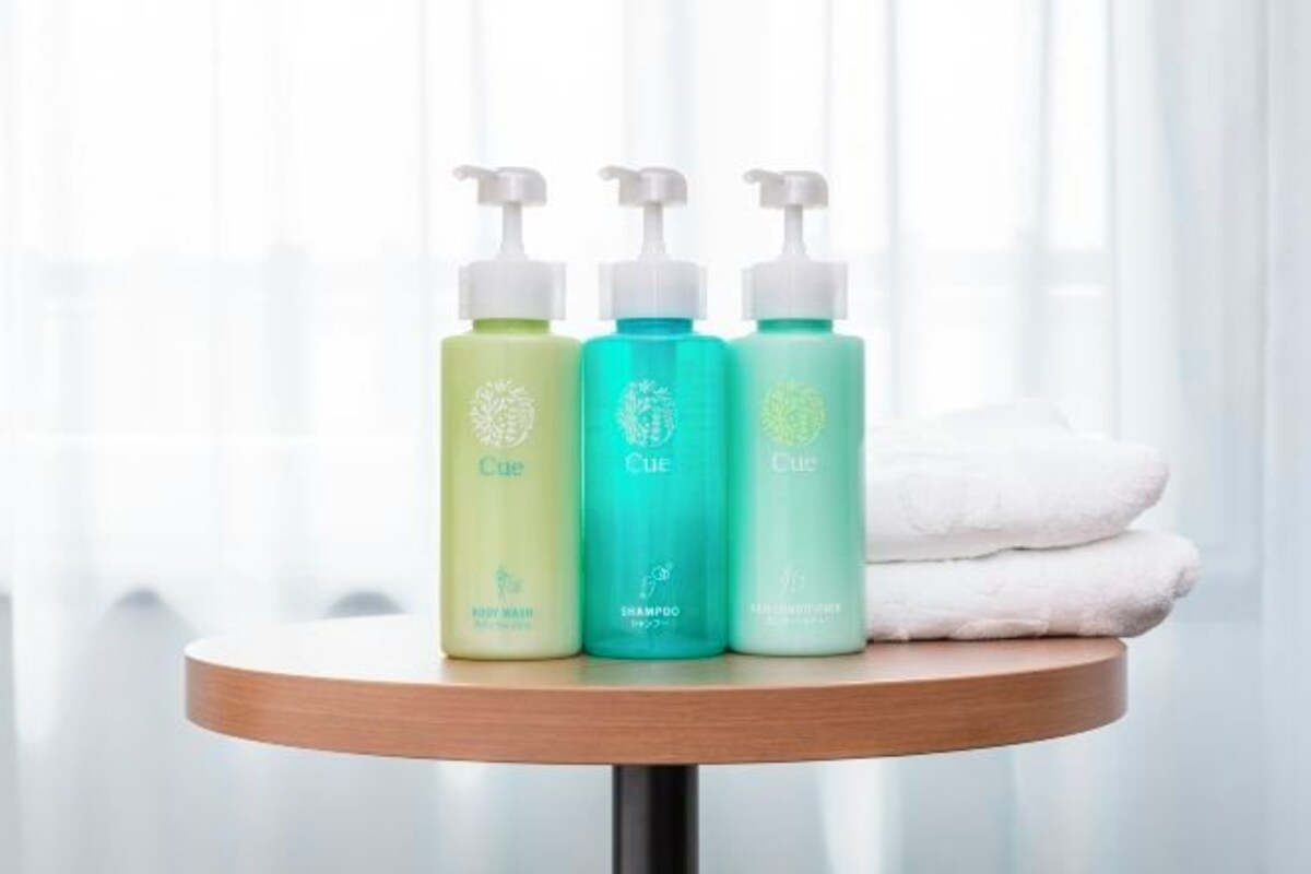 Three bottles of CUE body wash, shampoo, and conditioner on a wooden table with a white towel beside them.
