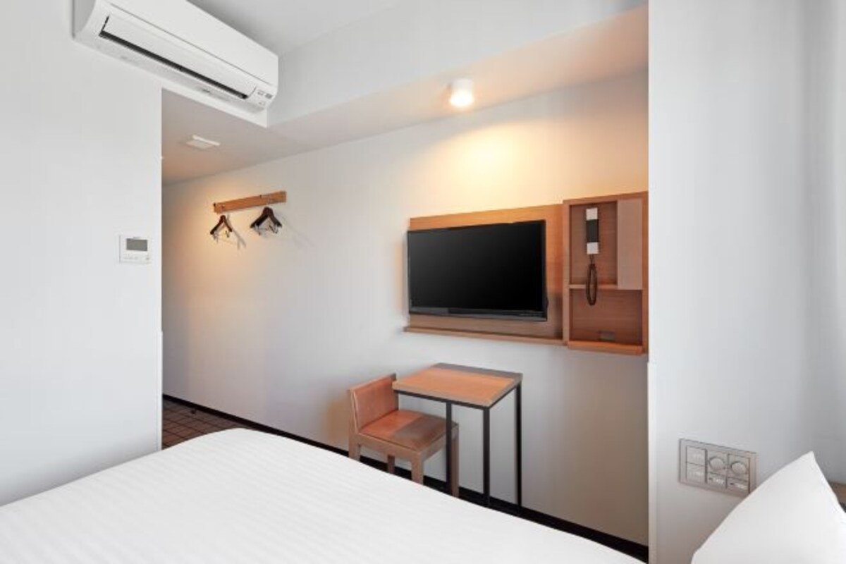 A hotel room with a bed, a desk with a chair, a flat-screen TV, and an air conditioning unit.