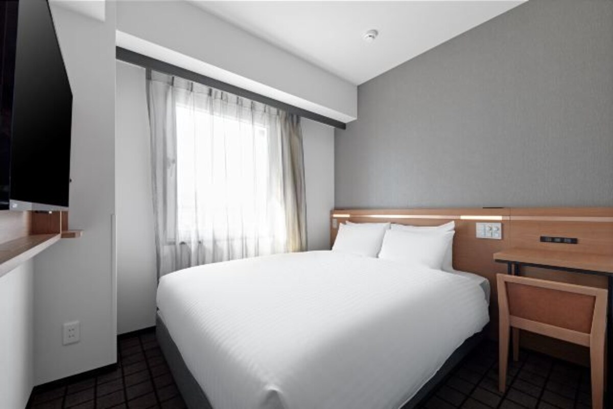 A hotel room with a large bed, a wooden headboard, a nightstand, a TV mounted on the wall, and a window with curtains.