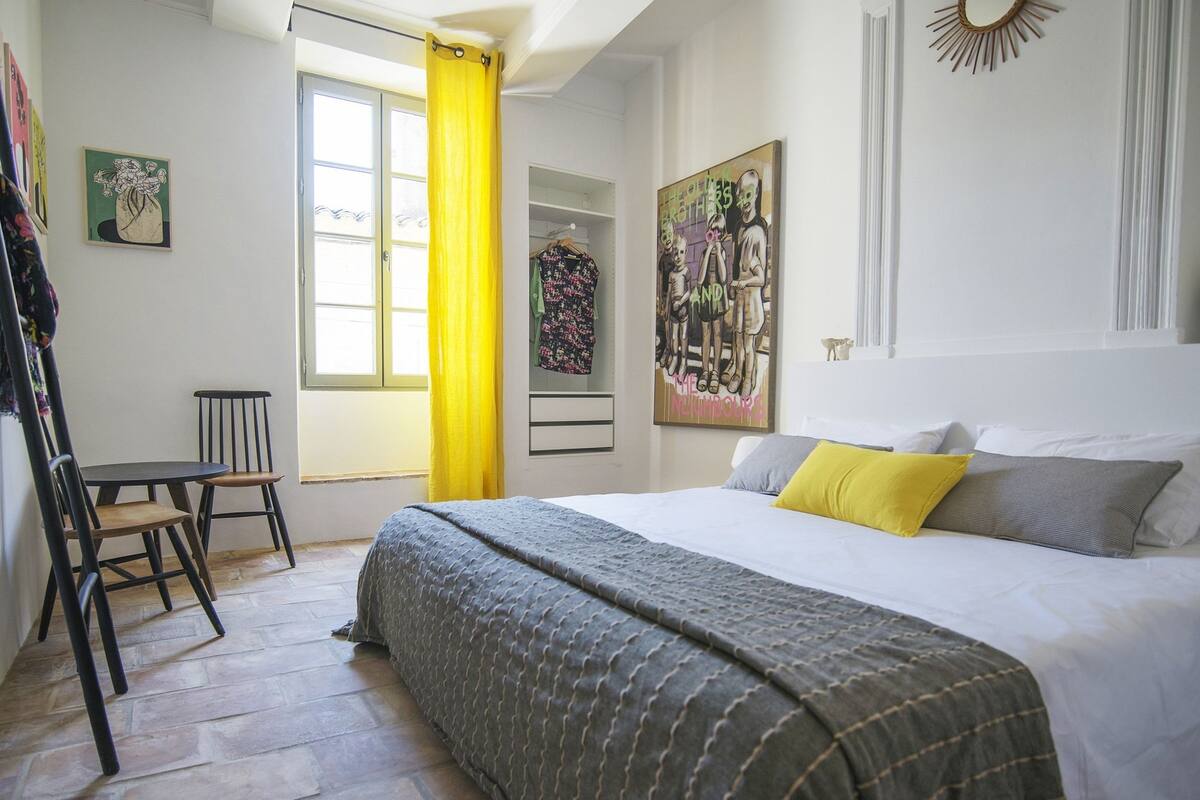 A bedroom with a bed, a chair, a window with yellow curtains, and artwork on the walls.