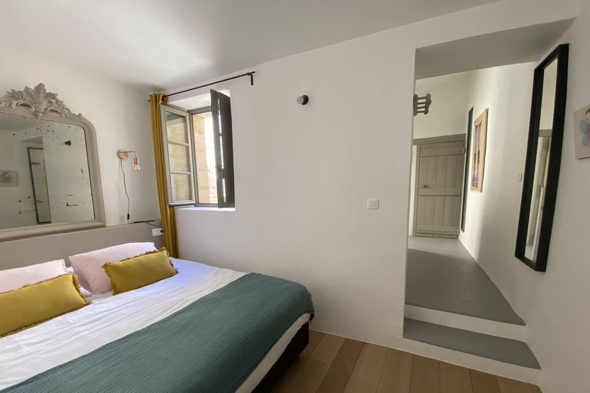 A bedroom with a bed, a mirror, a window with curtains, and a hallway.