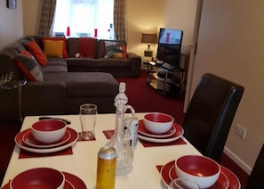 A dining area with a table set for four, a television in the background, and a sofa with cushions. - Huku Kwetu - Ravenhill House (Luton)