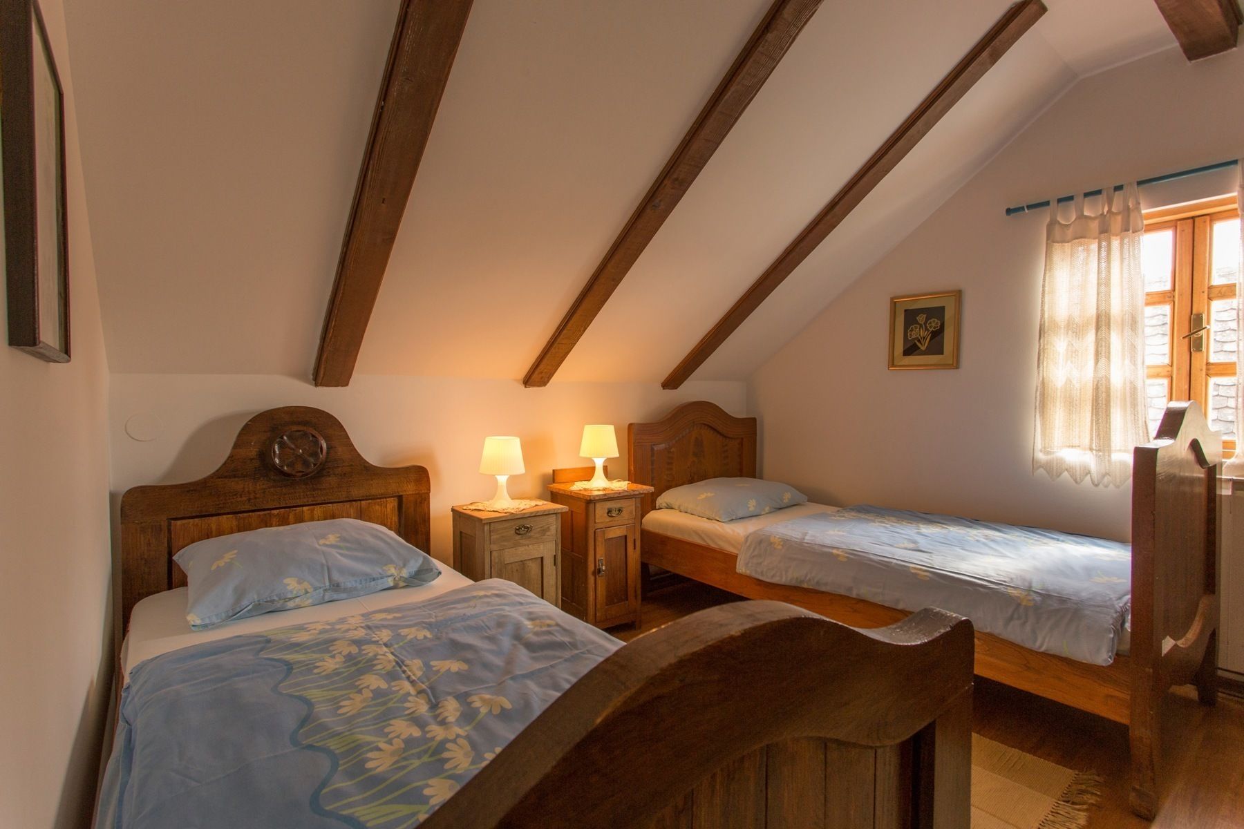 A bedroom with two wooden beds, a nightstand with lamps, a wooden chair, and a window with curtains.
