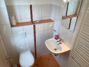 A bathroom with a white toilet, a sink with a mirror above it, and a shelf with toiletries. - Gästehaus Schulz (Heringsdorf)
