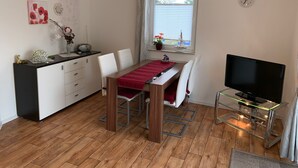 Ein kompakter Wohnbereich mit Esstisch, Stühlen, Fernseher, einer kleinen Küchenzeile und einem Fenster mit Jalousien.