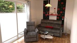 Ein Wohnzimmer mit grauem Sofa, grauem Sessel, kleinem weißen Couchtisch und einer Stehlampe. Ein Fenster bietet Blick auf eine grüne Umgebung.