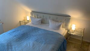 Ein Bett mit einer gesteppten blauen Decke, zwei weißen Kissen und einem gebürsteten Kopfteil. An beiden Seiten des Bettes befinden sich Nachttische mit Lampen.