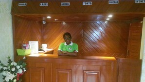 A reception desk with a wooden finish and a sign above listing destinations like Paris, Nairobi, Tokyo, Chicago, and Sydney. - Hôtel - Y (Douala)