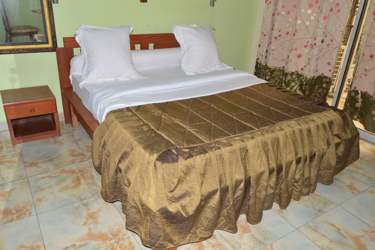 Una cama con colcha marrón, cabecera de madera y almohadas blancas. A la izquierda de la cama hay una mesita de noche con un cajón.