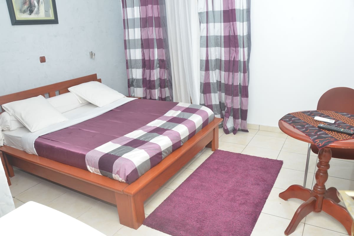 Un dormitorio con una cama de madera, una colcha de cuadros morada y blanca, una mesita redonda con mantel estampado y cortinas moradas.