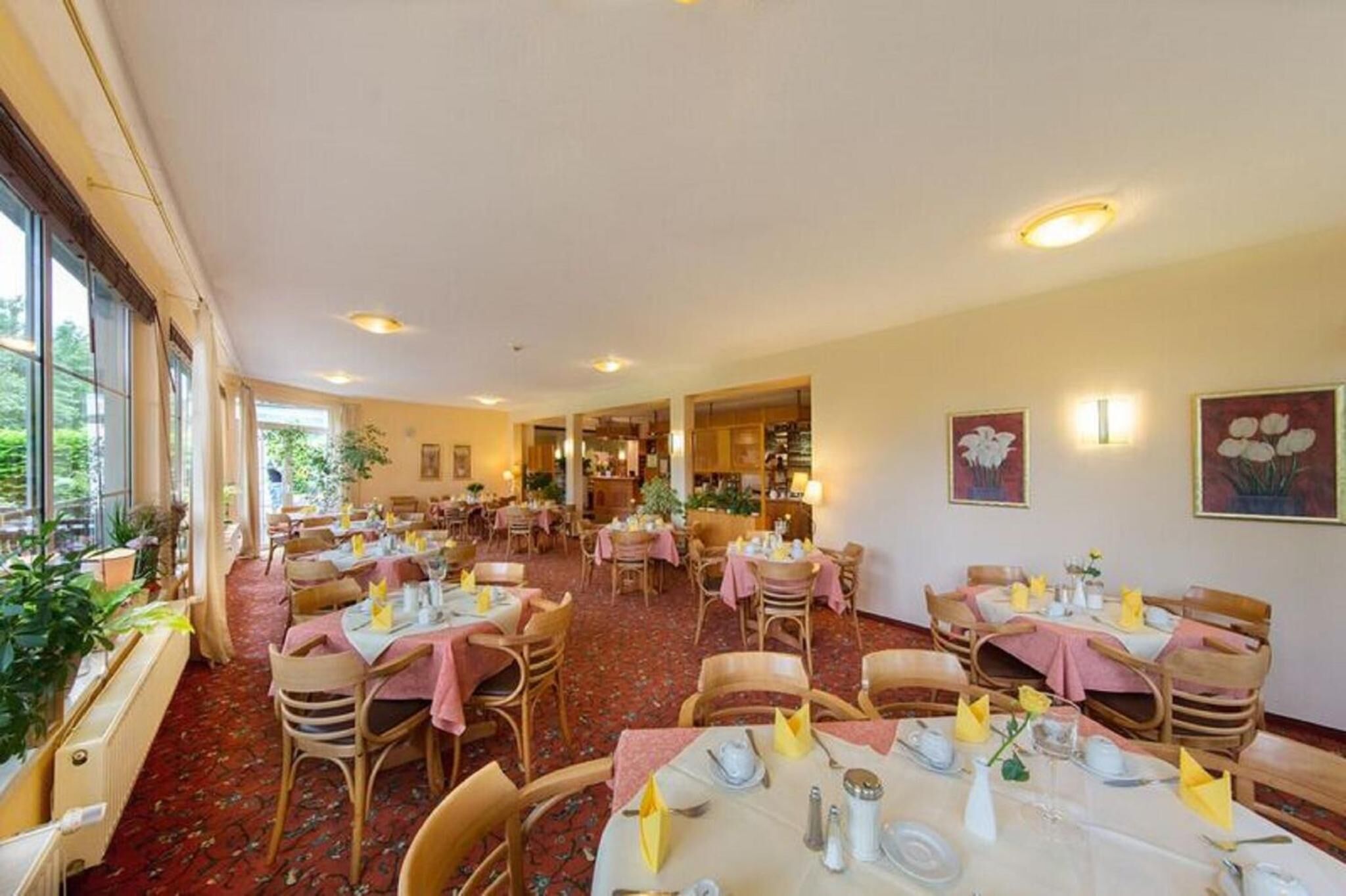 A spacious dining area with multiple tables set for dining, surrounded by large windows and potted plants.