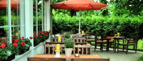 Un patio al aire libre con mesas y sillas de madera, sombrillas rojas y plantas en macetas.