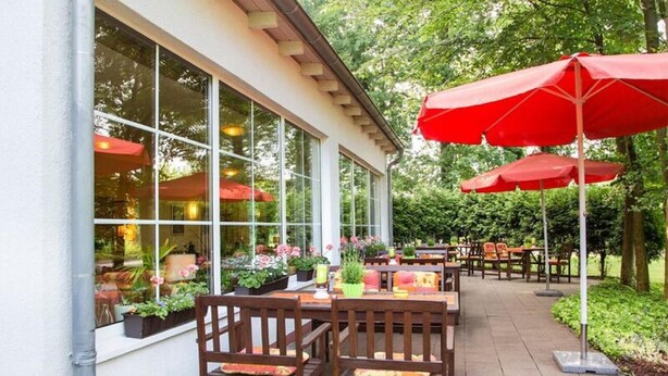 An outdoor patio with wooden tables and chairs, red umbrellas, and potted plants. - Montana Senden Münster (Senden)