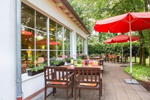 Un patio al aire libre con mesas y sillas de madera, sombrillas rojas y plantas en macetas.