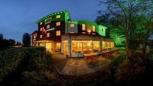 A hotel with a curved facade, large windows, and a well-lit outdoor area with tables and chairs. - Montana Senden Münster (Senden)