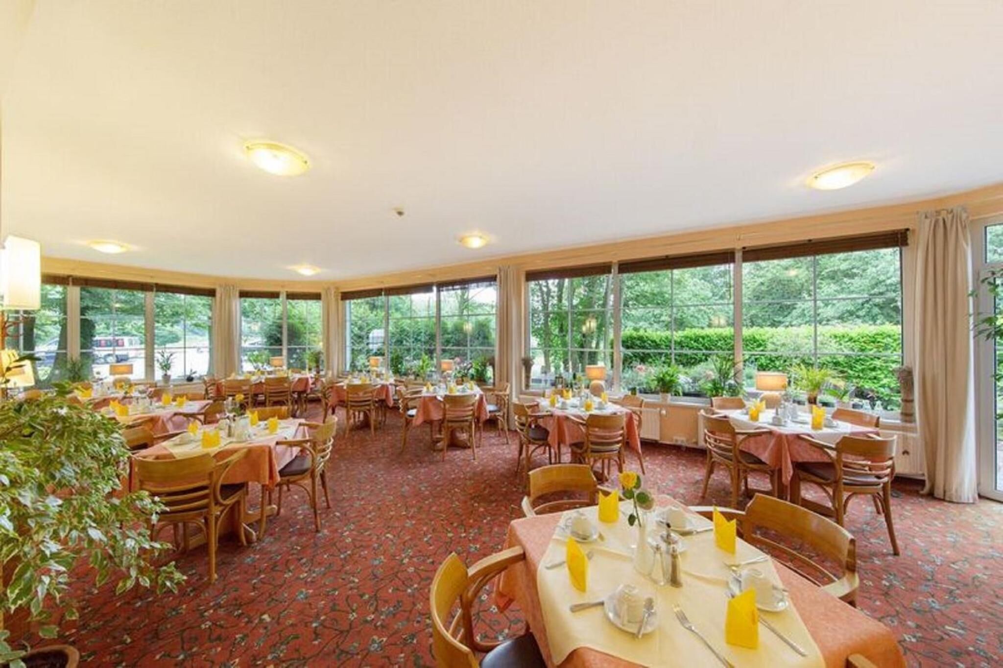 A spacious dining area with multiple tables and chairs, large windows offering a view of greenery outside.