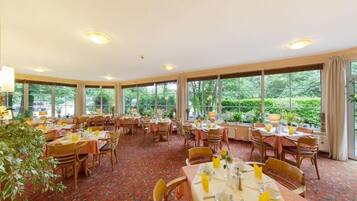 A spacious dining area with multiple tables and chairs, large windows offering a view of greenery outside.