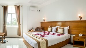 Habitación de hotel con cama de madera, un escritorio, una silla, una mesita de noche con lámpara y una ventana con cortinas.