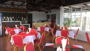 Un restaurante con varias mesas dispuestas con manteles blancos y rojos, sillas doradas y una zona de bar al fondo.