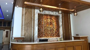 Una zona de recepción con un mostrador de madera, una pared de mármol con detalles tallados y un techo con paneles de madera e iluminación colgante.