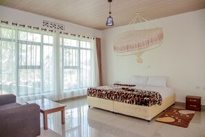 A bedroom with a bed, a nightstand, a sofa, a table, and large windows with curtains. - Musanze Caves Hotel (Muhoza)