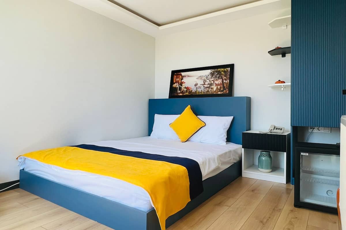 A modern hotel room with a bed, a bedside table, a small shelf, and a wall-mounted artwork.