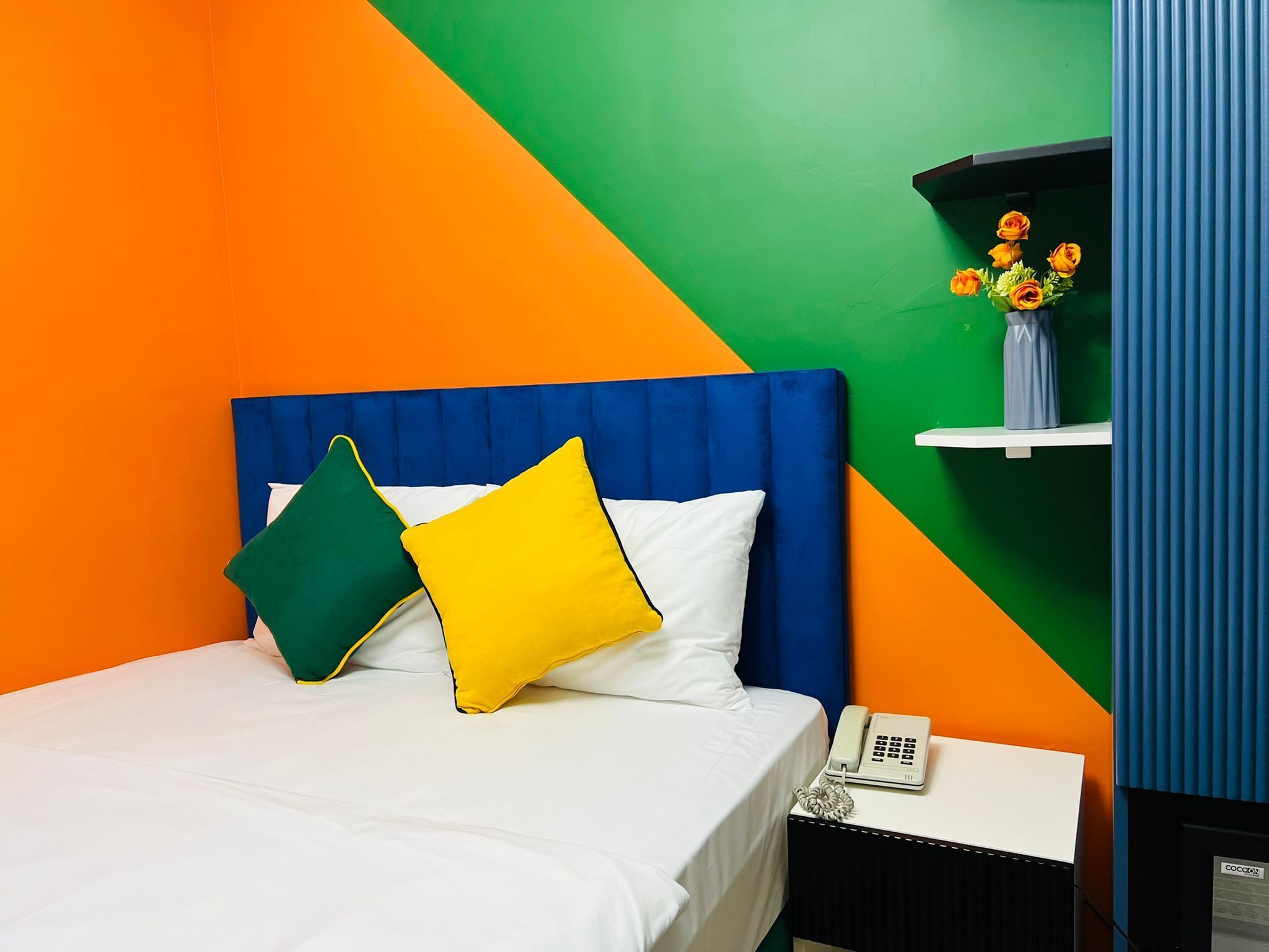 Une chambre d’hôtel avec un lit, une table de chevet avec un téléphone et une étagère avec des fleurs.