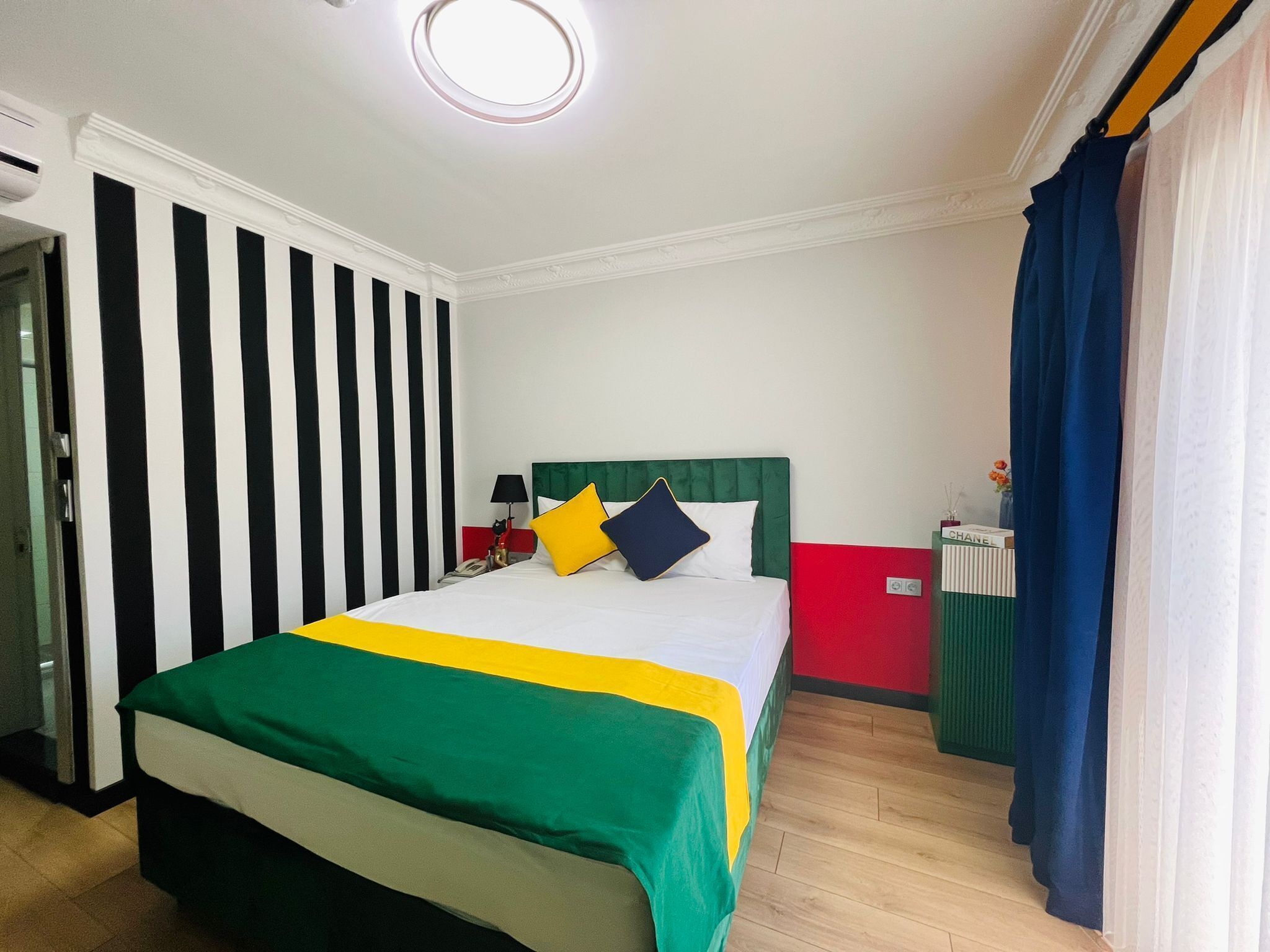 A hotel room with a bed, a nightstand, a lamp, a radiator, and a window with curtains.
