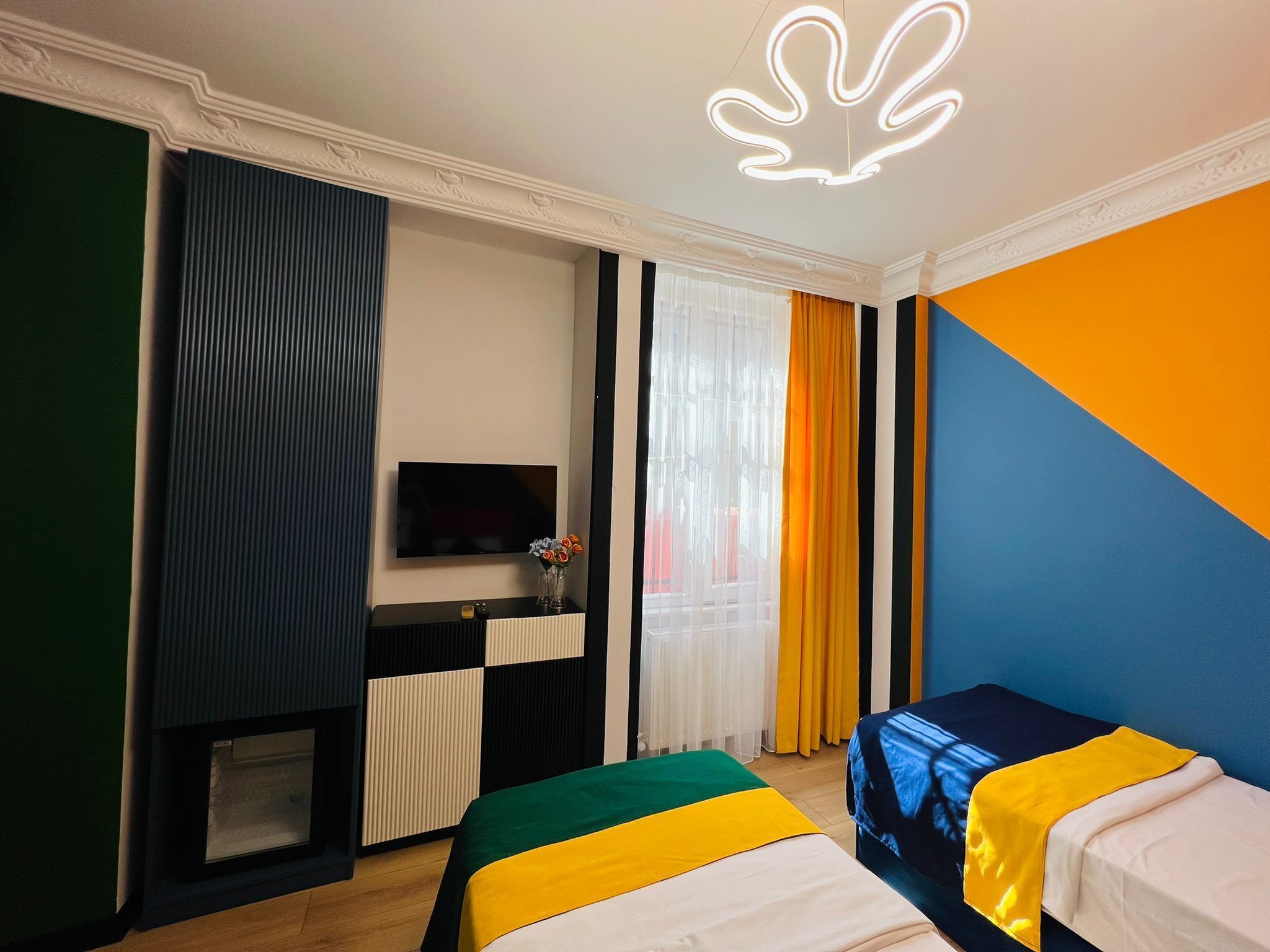A hotel room with a TV, a bed with a colorful bedspread, a small table with a vase, and a decorative ceiling light.