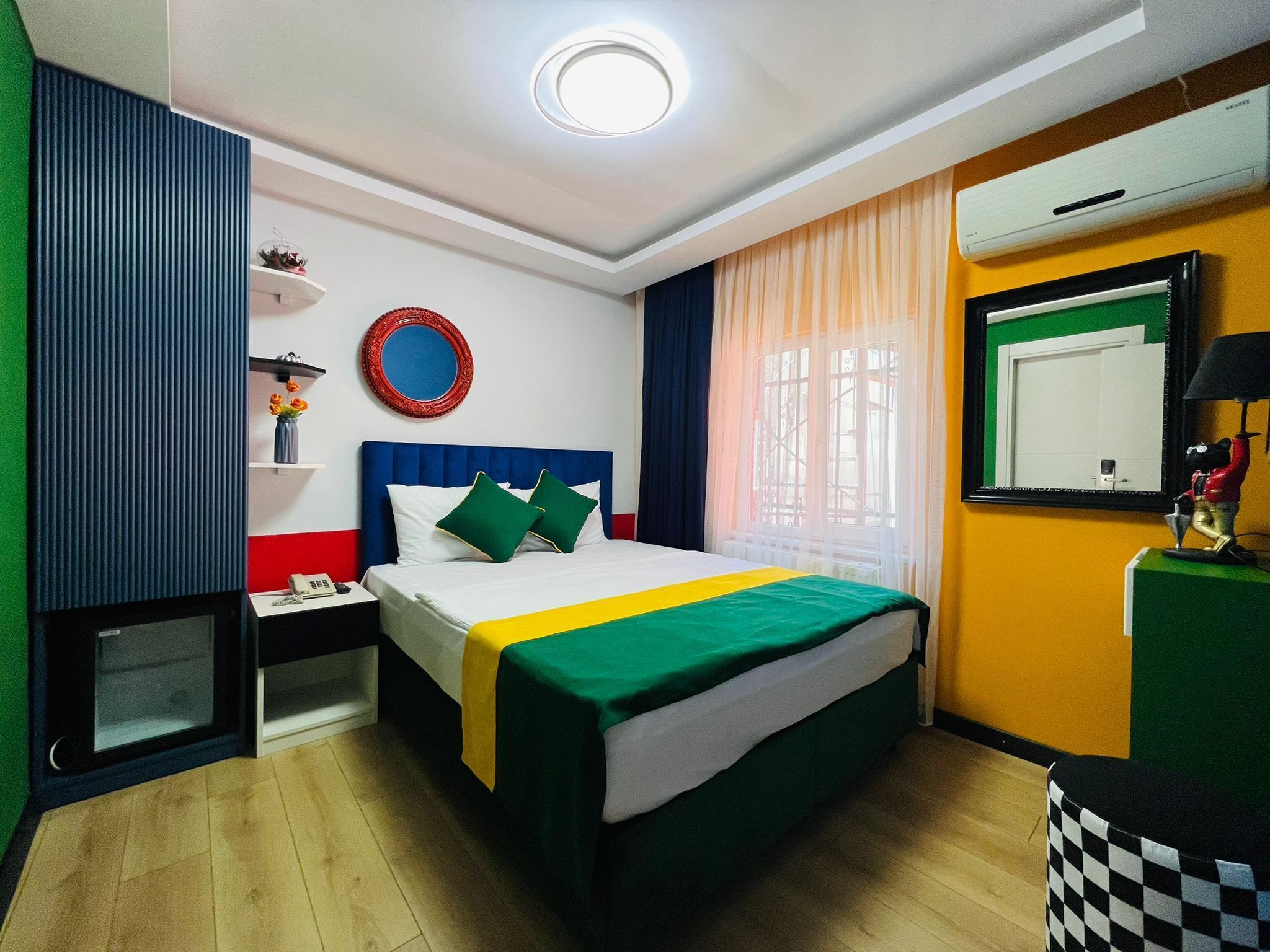 Ein Hotelzimmer mit Bett, Nachttisch, Spiegel, Fenster mit Vorhängen und einem kleinen Tisch mit Lampe.