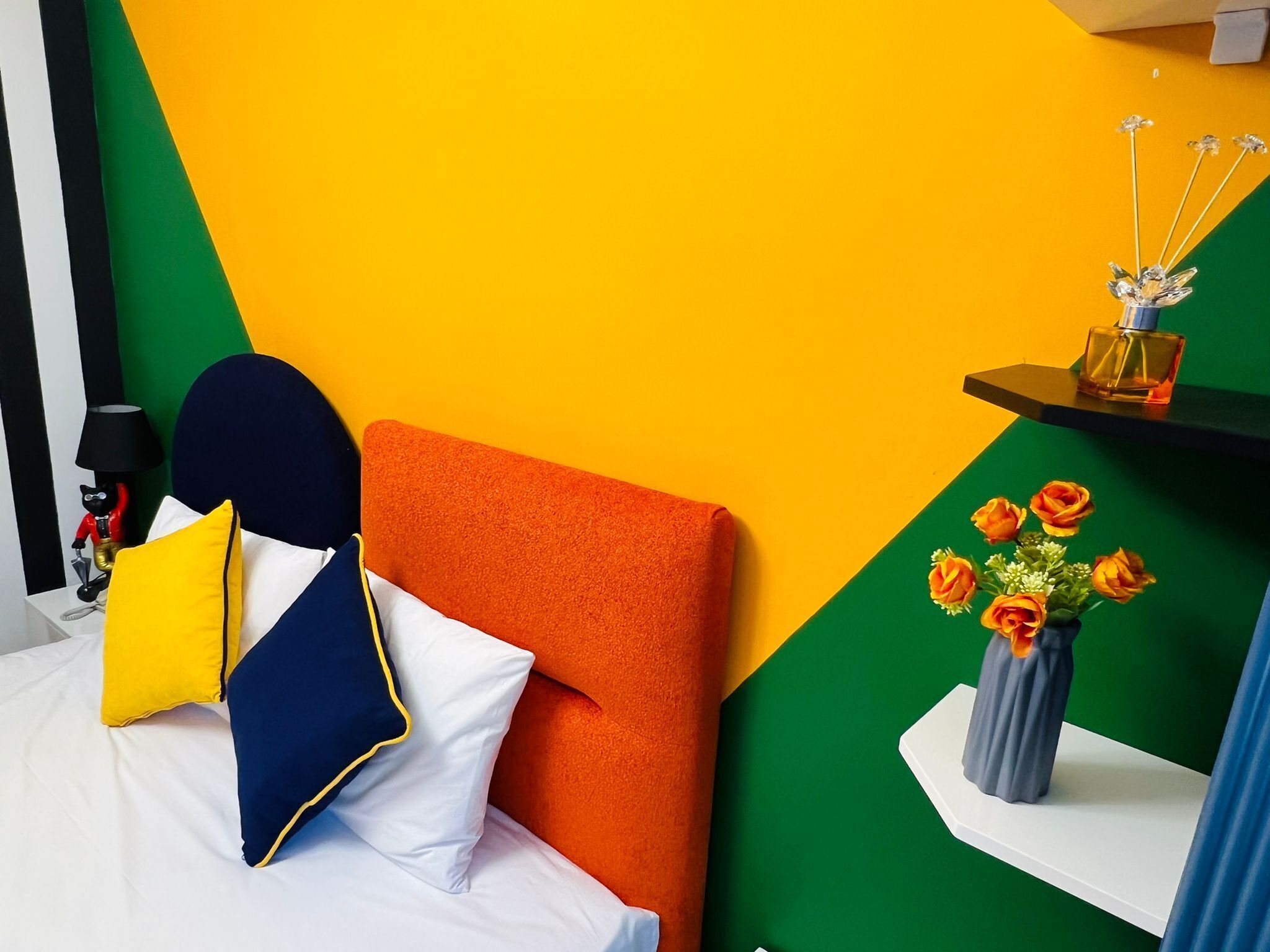 Una camera da letto moderna con un design geometrico vivace, un letto rifatto con cuscini colorati, un comodino con una lampada e un vaso con fiori arancioni.
