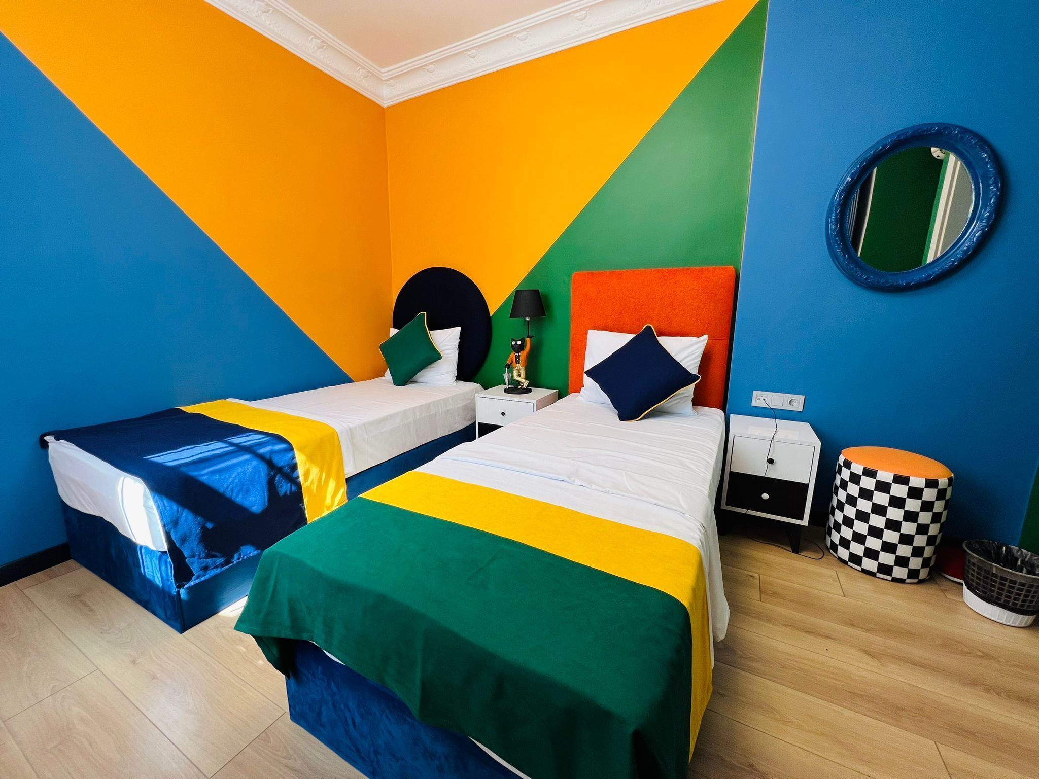 A room with two beds, a checkered ottoman, a mirror, and a nightstand with a lamp.